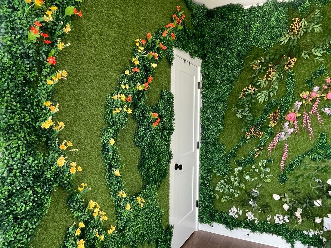 NC Office “Live” Wall: an office wall of green artificial foliage with pink, white and yellow flowers curving around a white door